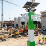 Auf einer Baustelle stehen grün-weiße Kameratürme mit Solarpanels, während Industrieanlagen und Baukräne in Betrieb sind.