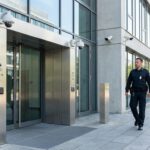 Vor einem modernen Bürogebäude mit Glasfassade kontrolliert ein Sicherheitsmitarbeiter den Eingang neben Überwachungskameras und Zutrittskontrolle.