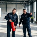 Zwei SecuConCept-Mitarbeiter besprechen in einer Halle die Brandwache und halten einen roten Feuerlöscher für den Einsatz bereit.