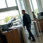 Mehrere Sicherheitskräfte kontrollieren einen Mann an einer hellen Eingangsschleuse, während draußen ein Polizeiwagen vor dem Glasgebäude steht.