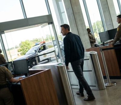 Mehrere Sicherheitskräfte kontrollieren einen Mann an einer hellen Eingangsschleuse, während draußen ein Polizeiwagen vor dem Glasgebäude steht.