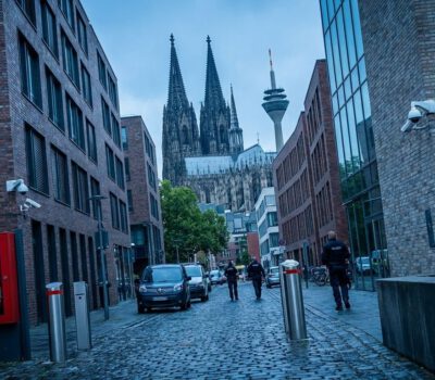 Vor dem Kölner Dom patrouillieren Sicherheitskräfte in einer engen Straße zwischen modernen Gebäuden und roten Wandhydranten.