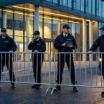 Vier Sicherheitskräfte in schwarzen Uniformen sichern bei Dämmerung einen Gebäudeeingang mit Absperrgittern und Fahrzeugen.